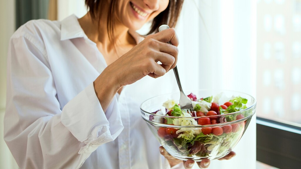 Woman mixing a fresh salad in a glass bowl near a window.