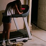 A tired person leaning over a treadmill in a dimly lit garage after a workout, with exercise equipment on the floor nearby.