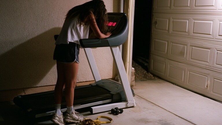 A tired person leaning over a treadmill in a dimly lit garage after a workout, with exercise equipment on the floor nearby.