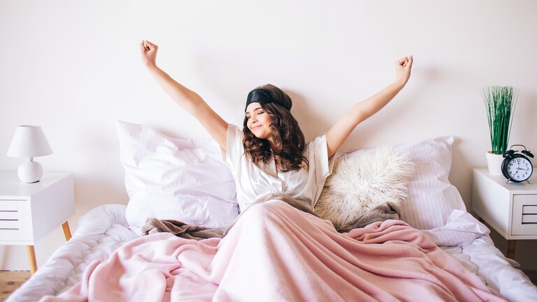 Woman stretching in bed after waking up, wearing a sleep mask, with white bedding, a bedside lamp, and an alarm clock nearby.