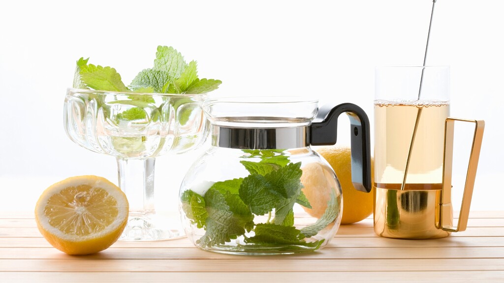 Herbal tea setup Lemon Balm leaves, a glass teapot, and a cup of pale tea on a light wooden table. 
