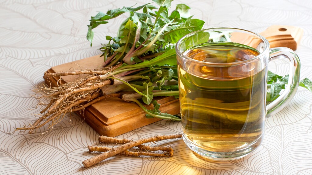 Cup of herbal tea beside fresh dandelion roots and greens on a wooden board.