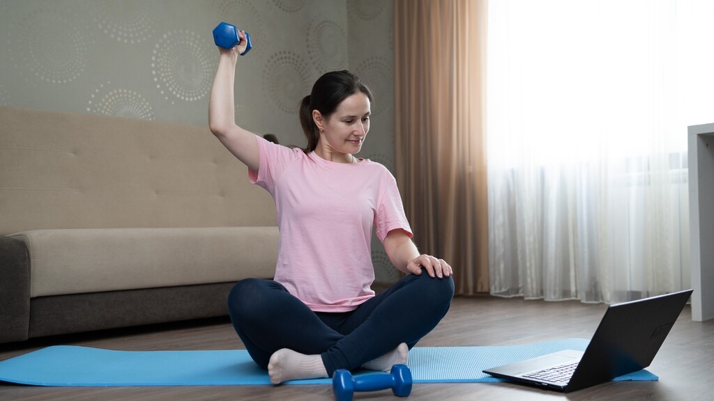 A woman sits on an exercise mat lifting a small dumbbell while following a laptop workout.
