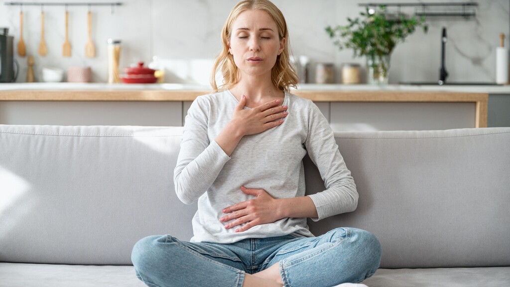 Woman sitting cross-legged on a couch with one hand on her chest and one on her stomach while taking a calming breath.
