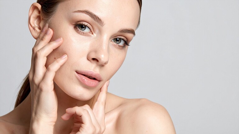 Woman with clear skin gently touching her face against a plain light background.