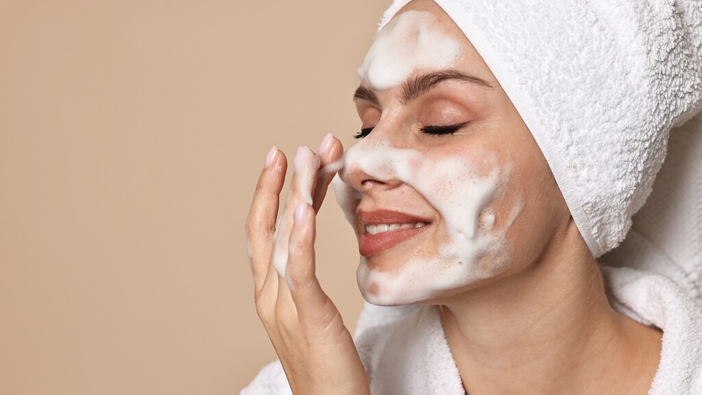Woman applying foaming cleanser to her face while wearing a white towel wrap.