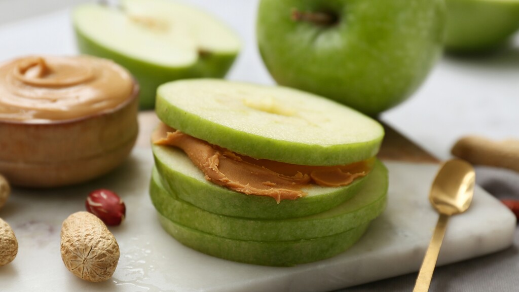 Green apple slices are layered with peanut butter on a cutting board.