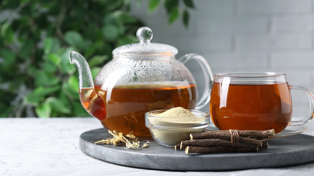 Herbal tea in a glass teapot and cup with licorice root sticks and powdered herb on a tray.