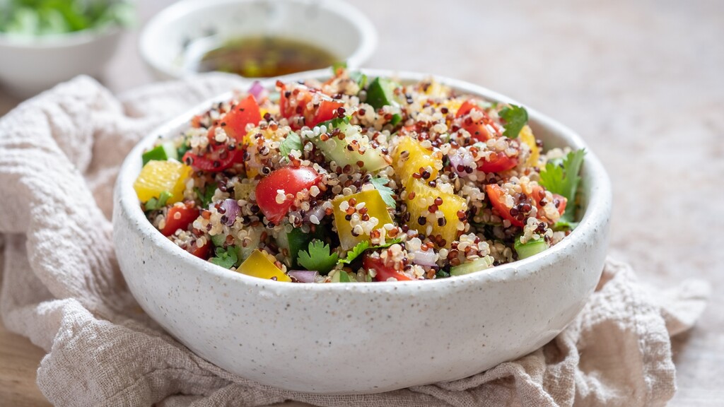 A white bowl filled with colorful quinoa salad, featuring tomatoes, cucumber, yellow bell pepper, red onion, and fresh herbs, with dressing in the background.