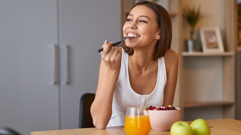 A woman smiling while eating a bowl of berries at a table with apples and a glass of juice.