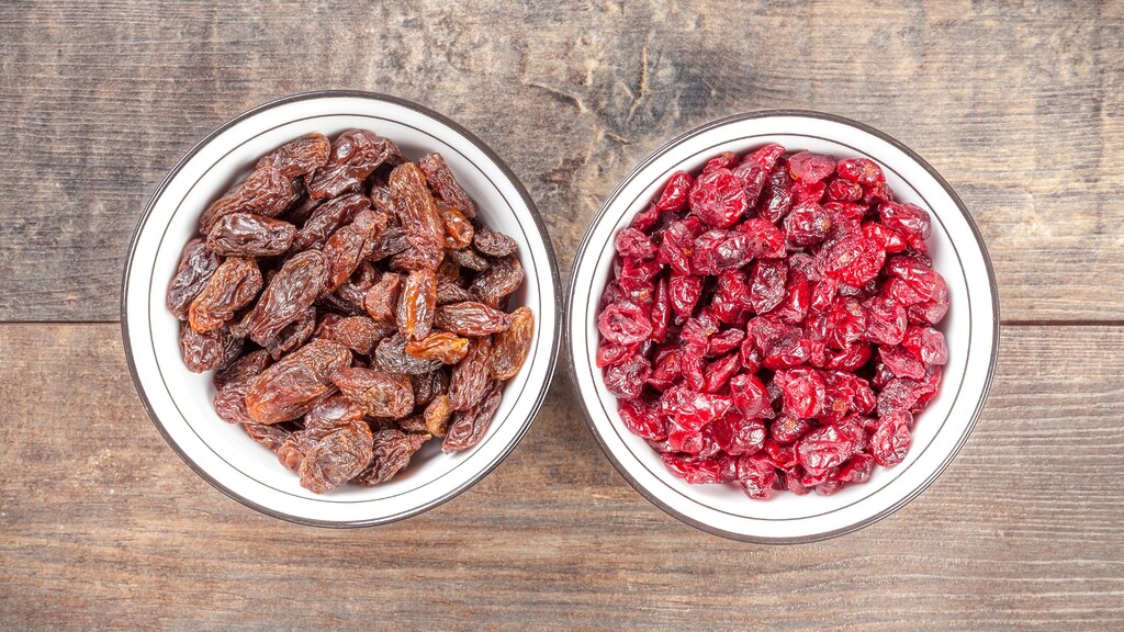 Two bowls of dried fruit, one filled with raisins and the other with dried cranberries, on a wooden table.