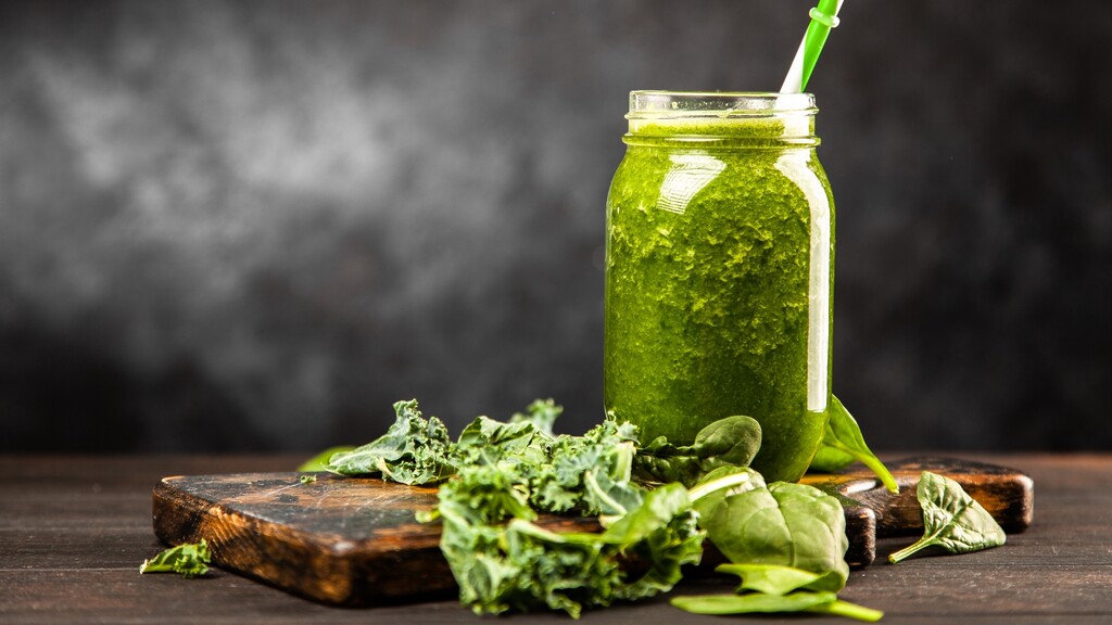 A green smoothie in a glass jar with kale and spinach leaves on a dark surface.