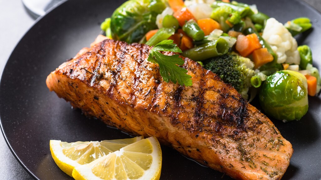 Grilled salmon served with mixed vegetables and lemon slices on a black plate.