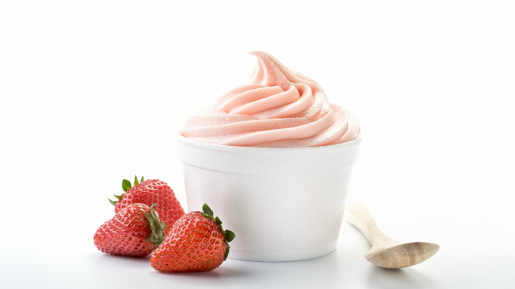 A cup of strawberry flavored yogurt with fresh strawberries and a wooden spoon.