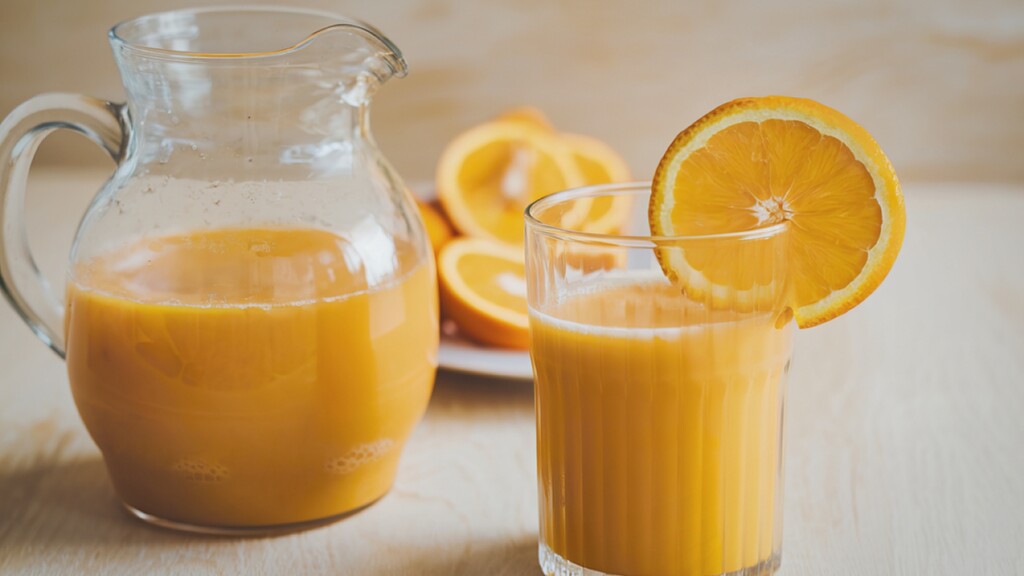 A glass and pitcher of orange juice with fresh orange slices in the background.