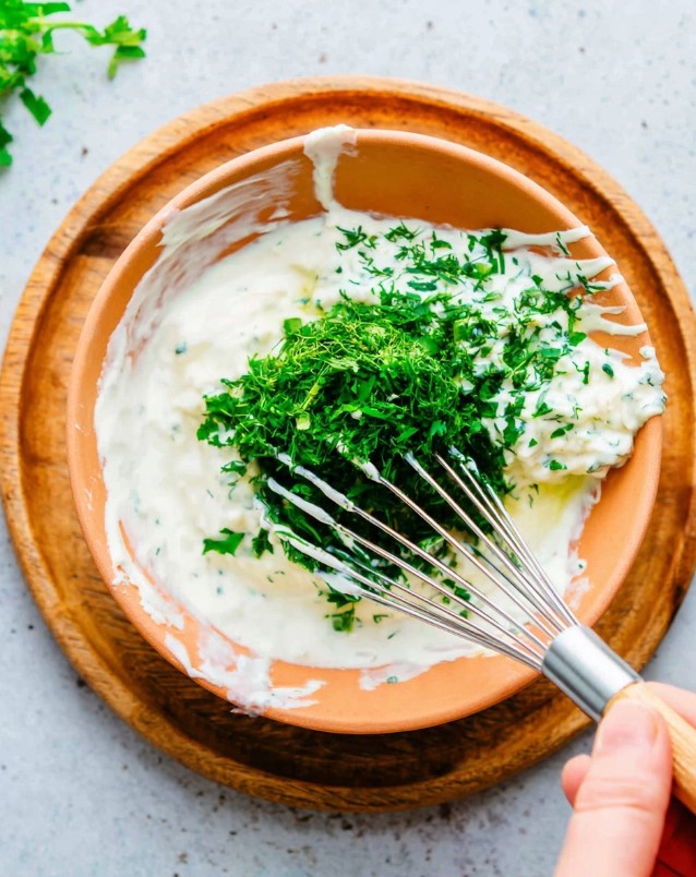 Minced herbs being whisked into the creamy white sauce base.
