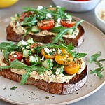 Thick sourdough loaded with creamy hummus, arugula, cherry tomatoes, cucumbers, and crumbled feta on a ceramic plate
