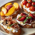Four slices of toasted sourdough topped with creamy ricotta, roasted cherry tomatoes, sautéed mushrooms, sliced peaches with prosciutto, and balsamic strawberries on a ceramic plate