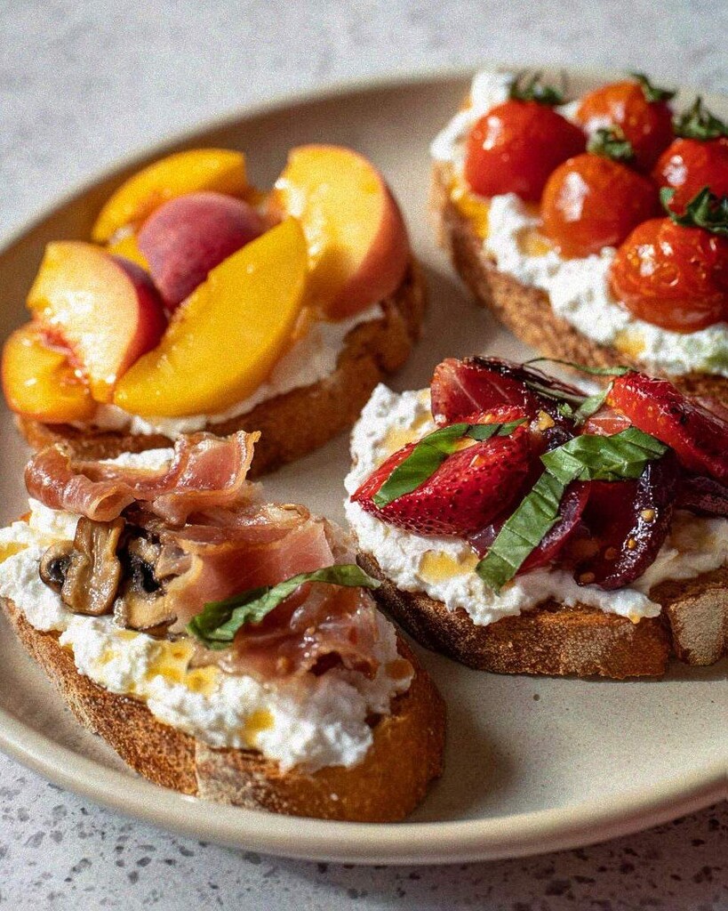 Four slices of toasted sourdough topped with creamy ricotta, roasted cherry tomatoes, sautéed mushrooms, sliced peaches with prosciutto, and balsamic strawberries on a ceramic plate
