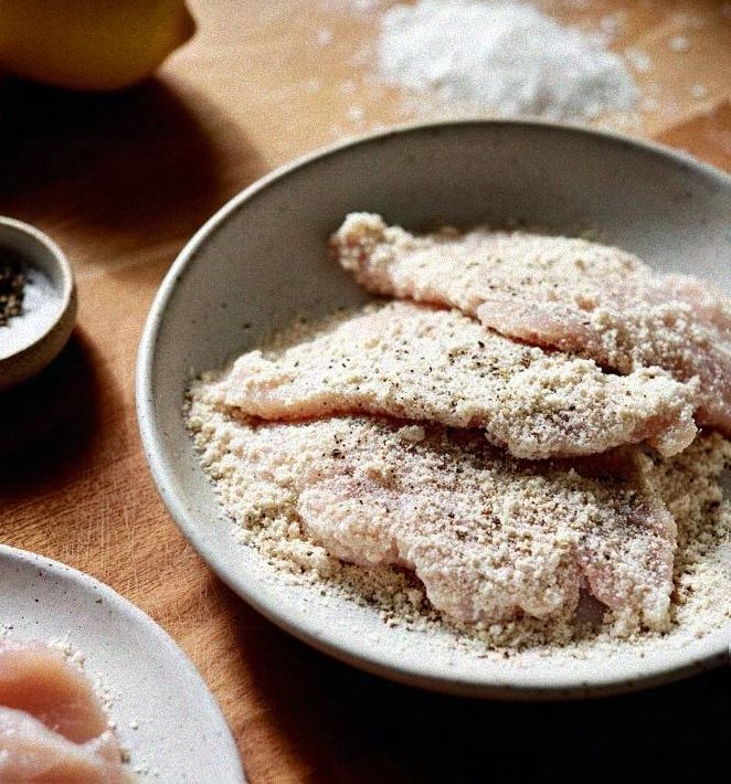 Thin chicken cutlets resting in a shallow bowl of seasoned whole wheat flour